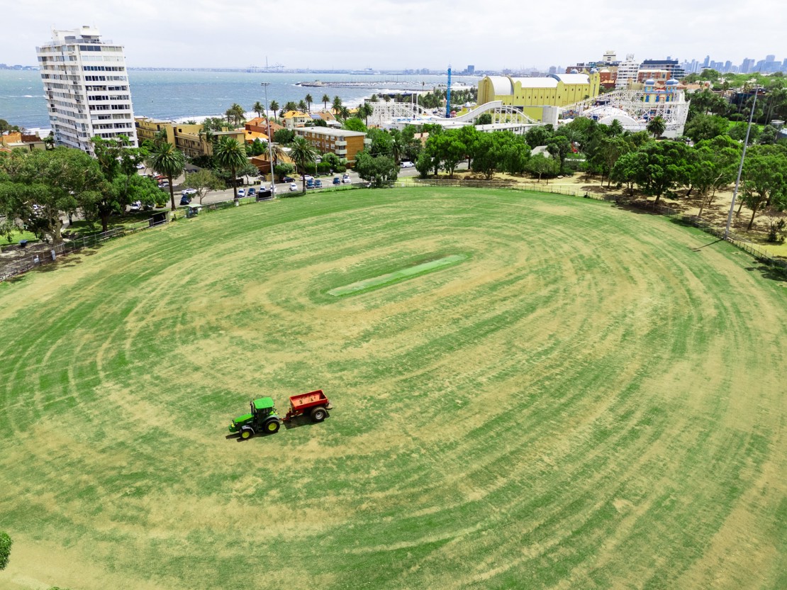Aerial view of a large oval sports field with a small green tractor towing a red trailer across the grass. Beyond the field are trees, residential buildings and a coastal foreshore, with a roller coaster and amusement park beside the bay under an overcast sky.