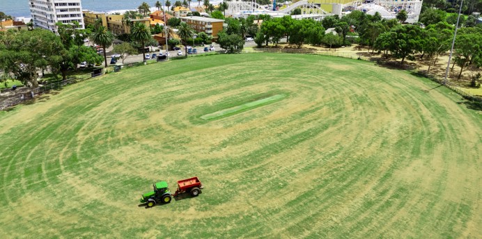 Aerial view of a large oval sports field with a small green tractor towing a red trailer across the grass. Beyond the field are trees, residential buildings and a coastal foreshore, with a roller coaster and amusement park beside the bay under an overcast sky.
