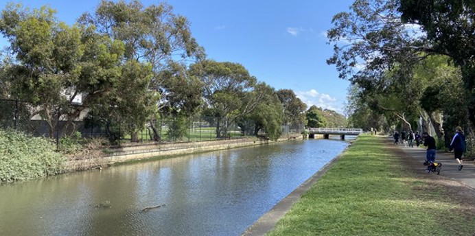 A calm canal lined with trees on both sides under a clear blue sky. A grassy path runs alongside the water, where several people are walking in the distance. A small bridge crosses the canal ahead, and the water reflects the bright midday sunlight.