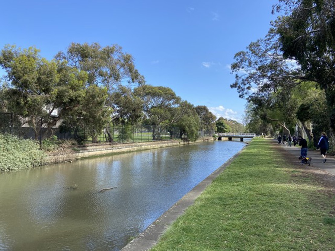 A calm canal lined with trees on both sides under a clear blue sky. A grassy path runs alongside the water, where several people are walking in the distance. A small bridge crosses the canal ahead, and the water reflects the bright midday sunlight.