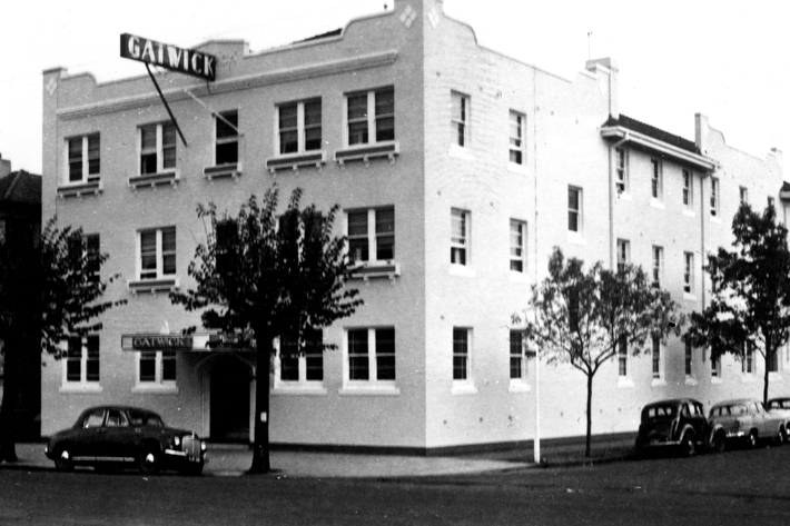 Street view of the Gatwick hotel on a corner block, trees and parked vintage cars in front of it.