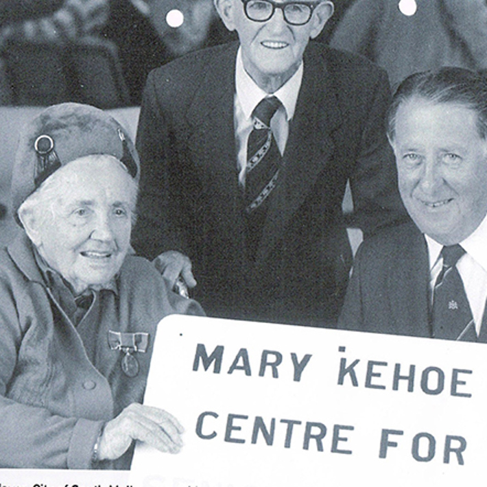 Mary Kehoe sitting at a table holding the sign for the community centre named in her honour. Seated with the mayor of South Melbourne Bert Jones. Mary's husband, Jack, is standing behind them.