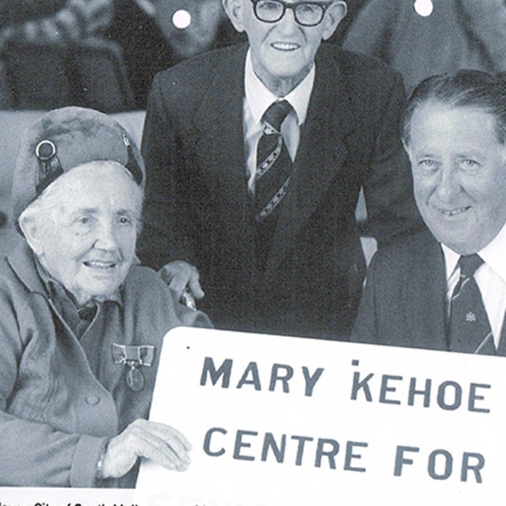 Mary Kehoe sitting at a table holding the sign for the community centre named in her honour. Seated with the mayor of South Melbourne Bert Jones. Mary's husband, Jack, is standing behind them. 