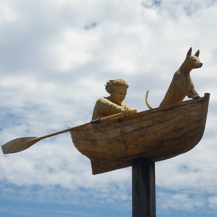 Sculpture of a boat with a person rowing and a dog riding up front raised off the ground at the entrance to Gasworks Arts Park off Graham Street