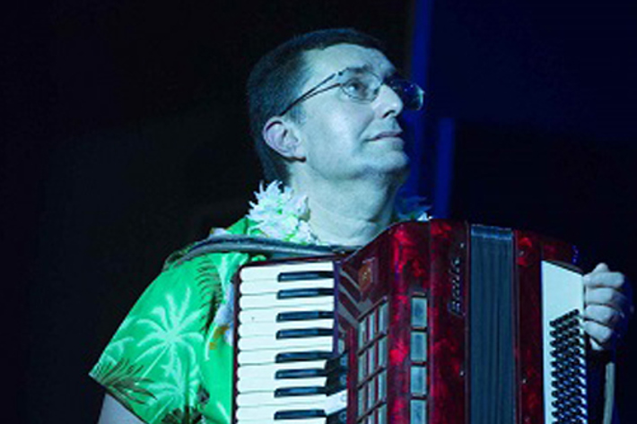 A sole performer wearing a green shirt holding a red piano accordian stares up with a sweet melancholy look on his face