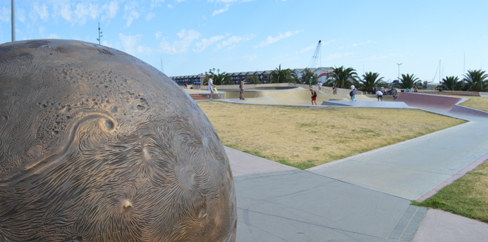 Skaters using the Marina Skate Park with the boat sheds in the background. Wide paved footpaths provide easy access.  