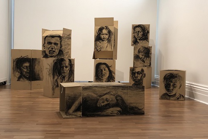 A wooden floored white walled gallery space containd brown carboard boxes of various sizes on which the artist has drawn very realistic faces in black wan portraits