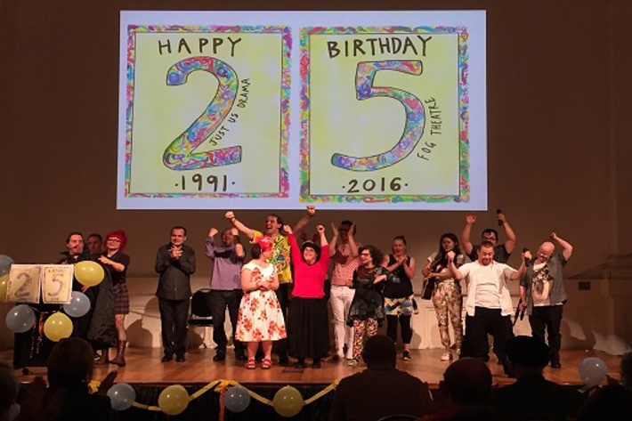 A group of performers in a party like atmosphere gathered together onsatge in front of two placards that say Happy Birthday 25 years