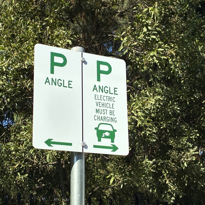 A photo of a parking sign. The left sign is unrestricted parking, the right sign is for charging EVs only and has text that reads "electric vehicle must be charging".