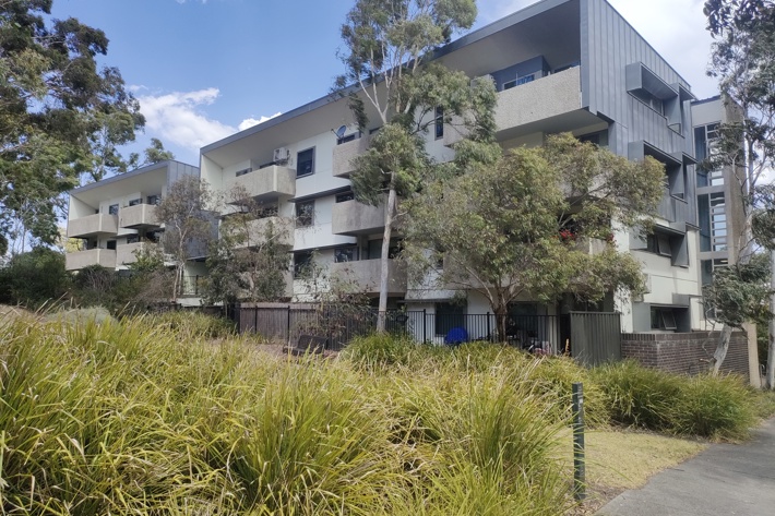 A photo of a medium-rise apartment building, surrounded by green trees and shrubs.