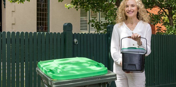 A woman stands next to a FOGO bin, she's holding a kitchen caddy 