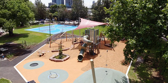 Elevated view of a playground with tan rubber flooring, climbing structures, slides, and a large pink shade sail. The playground is surrounded by trees and green grass, with a blue basketball court visible in the background. Tall buildings can be seen in the distance beyond the park.