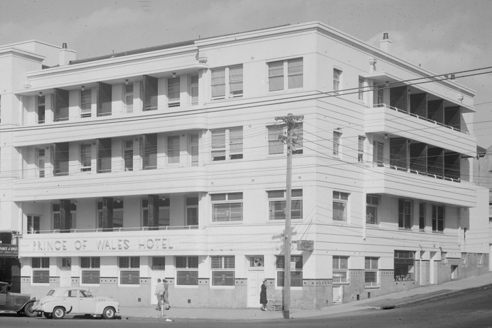 street view looking up at four storey art deco style hotel on a corner block balconies run the length of the building on each floor.